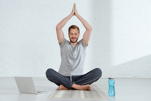 young man practicing half lotus pose with raised prayer hands near laptop and sports bottle - Foto, afbeelding