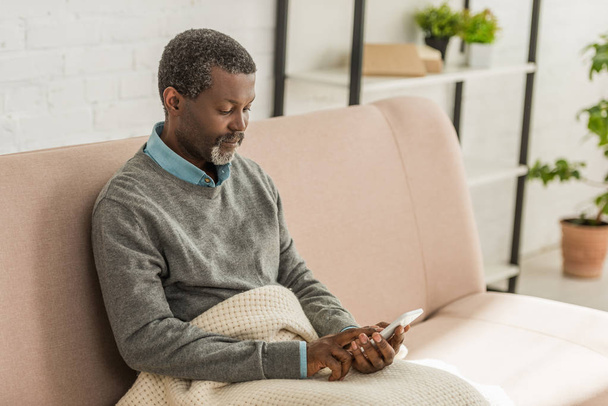 przemyślany Afrykanin amerykański człowiek siedzi na kanapie i za pomocą smartfona - Zdjęcie, obraz