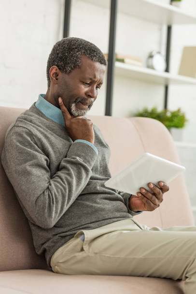 uomo afroamericano premuroso seduto sul divano e utilizzando tablet digitale
 - Foto, immagini