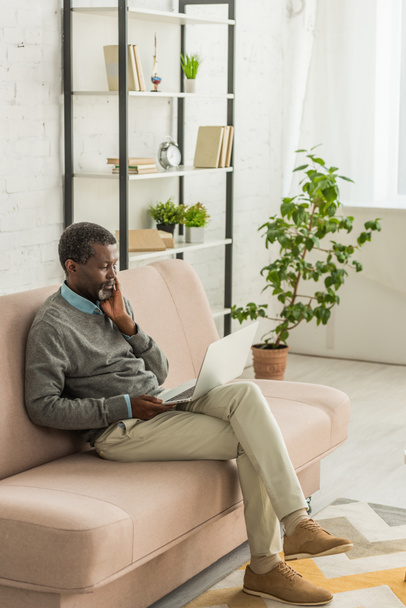 thoughtful african american man sitting on sofa in living room and using laptop - Фото, зображення