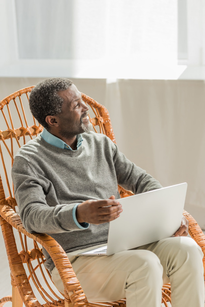 alegre afroamericano hombre sentado en silla de mimbre con cuaderno y mirando hacia otro lado
 - Foto, Imagen