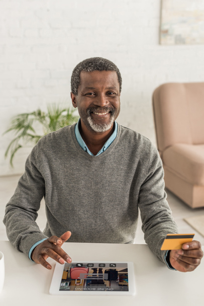 vidám afro-amerikai férfi kezében hitelkártya és digitális tablet használata foglalási alkalmazás a képernyőn - Fotó, kép
