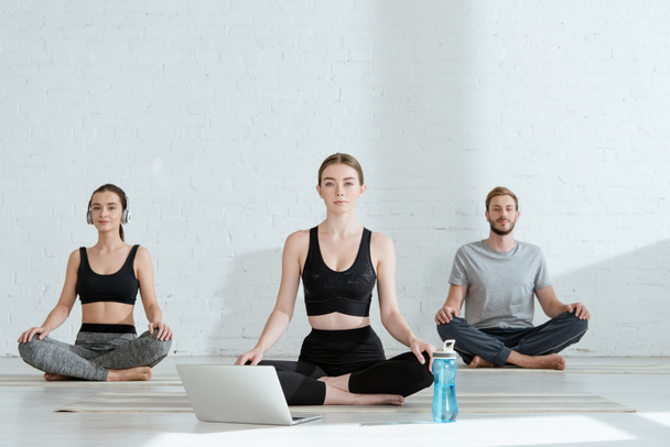 jonge mannen en vrouwen die yoga beoefenen in de helft lotus poseren in de buurt laptop en sportfles - Foto, afbeelding