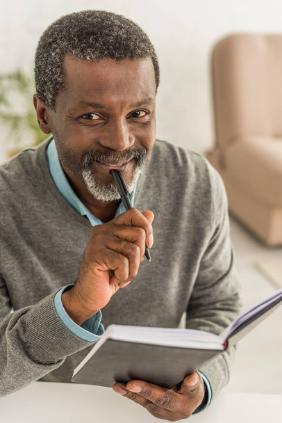  vidám afro-amerikai férfi kezében toll és notebook miközben mosolyog a kamera - Fotó, kép
