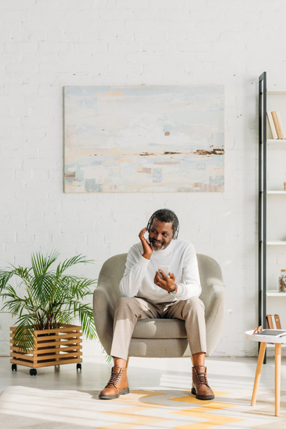 κομψό αφροαμερικανός άνδρας κάθεται στον καναπέ στο σαλόνι και ακούγοντας μουσική στα ακουστικά - Φωτογραφία, εικόνα