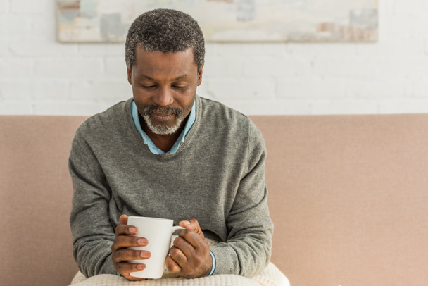 älterer afrikanisch-amerikanischer Mann sitzt mit Decke auf Knien und hält Tasse mit wärmendem Getränk - Foto, Bild