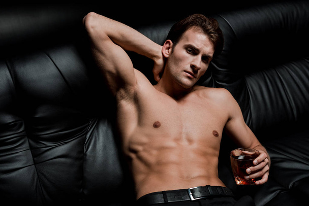 handsome shirtless man sitting on sofa with glass of whiskey in dark room - Fotó, kép