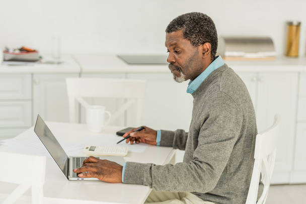 σοβαρός Αφροαμερικάνος που χρησιμοποιεί φορητό υπολογιστή κατά τον υπολογισμό των εξόδων - Φωτογραφία, εικόνα