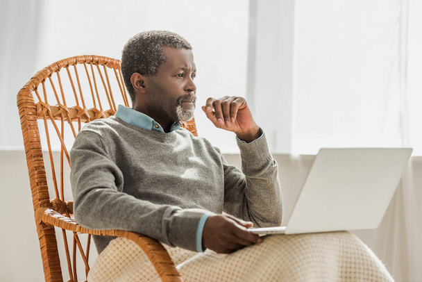 verträumter afrikanisch-amerikanischer Mann sitzt im Korbstuhl mit Laptop und schaut weg - Foto, Bild