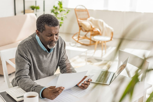 enfoque selectivo del hombre afroamericano senior mirando la factura de servicios públicos mientras está sentado cerca de la computadora portátil
 - Foto, imagen