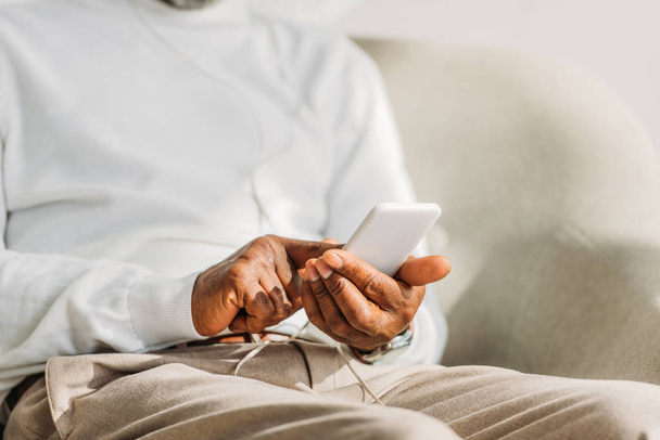 cropped view of african american man in white pullover sitting and using smartphone  - Valokuva, kuva