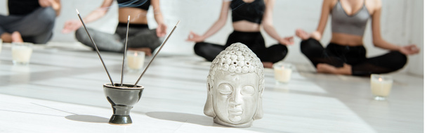 selektiver Fokus von dekorativem Buddhakopf, aromatischen Stöcken und Kerzen und jungen Leuten, die Yoga in halber Lotus-Pose praktizieren, Panoramaaufnahme - Foto, Bild