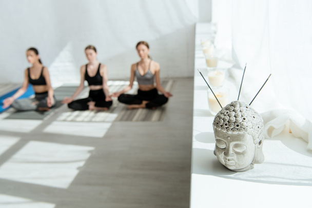 selective focus of Buddha head sculpture with aromatic sticks on windowsill, and three women sitting in half lotus pose - Photo, Image