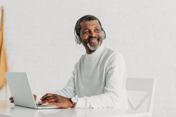americano africano alegre em fones de ouvido usando laptop e olhando para longe
 - Foto, Imagem
