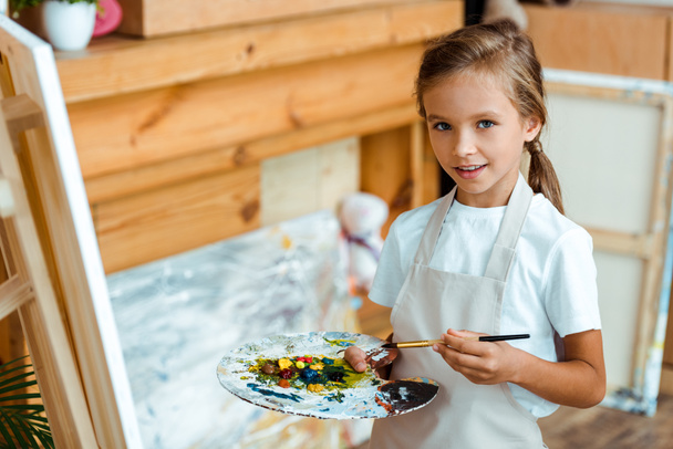happy kid in apron holding palette with colorful gouache paint  - Photo, Image