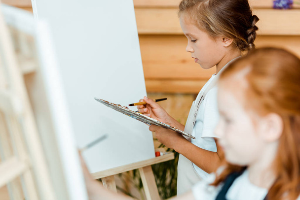 selective focus of cute kid holding palette near easel and child  - Photo, Image