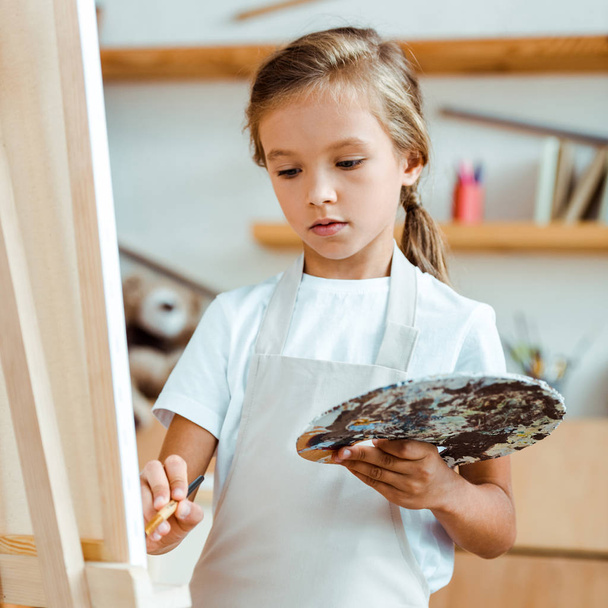 selective focus of kid in apron holding palette with colorful gouache paint and paintbrush - Foto, afbeelding