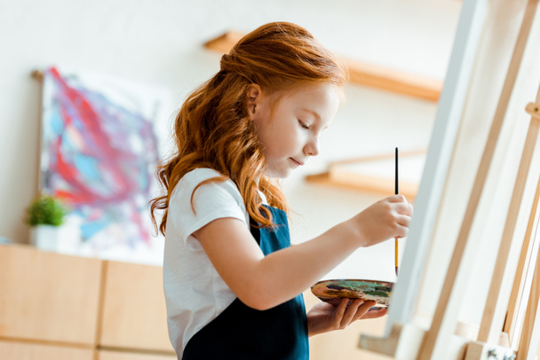 selective focus of cute redhead kid holding palette and paintbrush near easel  - Φωτογραφία, εικόνα