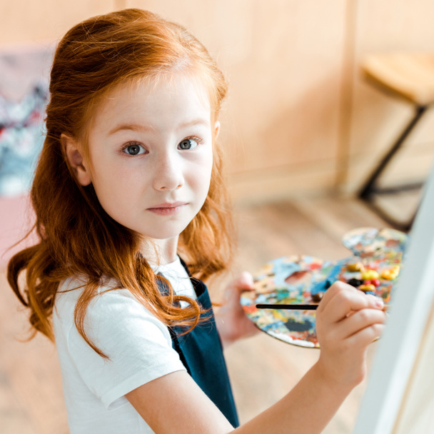 selective focus of cute redhead child holding paintbrush and looking at camera  - Фото, зображення