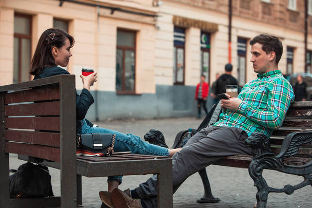 Смеющийся мужчина с женщиной, сидящей на скамейке и пьющей кофе-серфинг в интернете. городской образ жизни
 - Фото, изображение