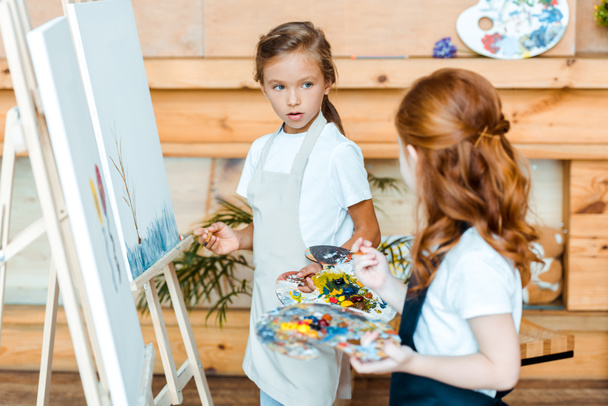 selective focus of adorable kid looking at redhead child near easels in art school - Photo, Image