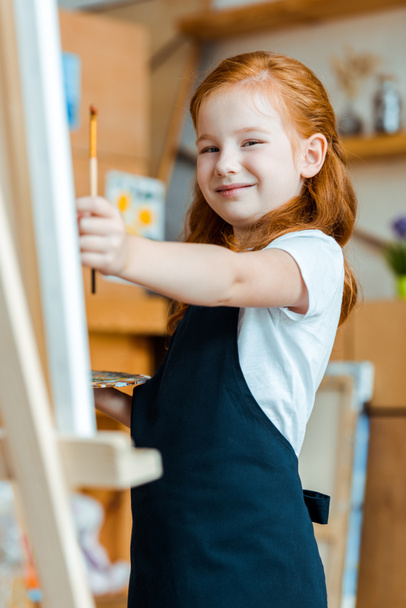selective focus of positive redhead child holding paintbrush  - Φωτογραφία, εικόνα