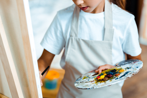 cropped view of kid with palette standing near easel  - 写真・画像