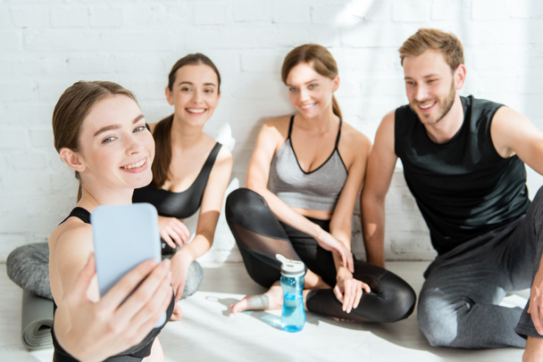 cheerful girl taking selfie on smartphone with friends sitting in easy poses - Photo, Image