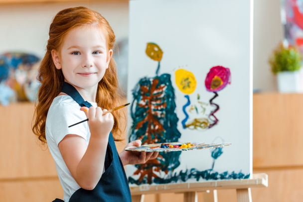 sonriente pelirroja niño de pie cerca de pintura sobre lienzo en la escuela de arte
  - Foto, Imagen