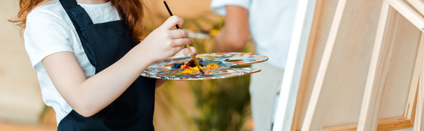 panoramic shot of kid holding palette and paintbrush near easel  - Foto, Bild