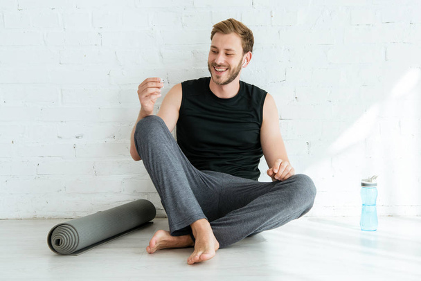 hombre alegre sentado en el suelo cerca de la esterilla de yoga y la botella de deportes y la celebración de auriculares
 - Foto, imagen