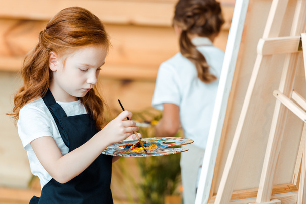 selektivní zaměření roztomilé zrzky dítě malování v umělecké škole v blízkosti dítěte - Fotografie, Obrázek