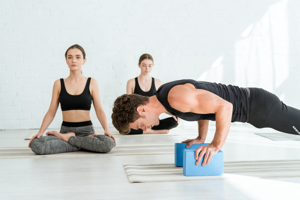 young man practicing low push up plank pose on stands, and women sitting in half lotus pose - Foto, Bild
