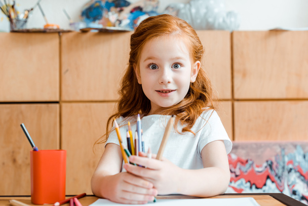 niño pelirrojo excitado sosteniendo lápices de color en la escuela de arte
  - Foto, Imagen