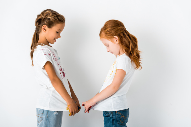 side view of cheerful kids with paint on t-shirts standing on white  - Foto, Imagen