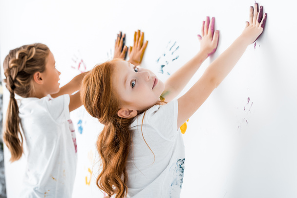 επιλεκτική εστίαση ευτυχισμένη κοκκινομάλλα παιδί βάζοντας τα αποτυπώματα των χεριών κοντά στο παιδί σε λευκό  - Φωτογραφία, εικόνα