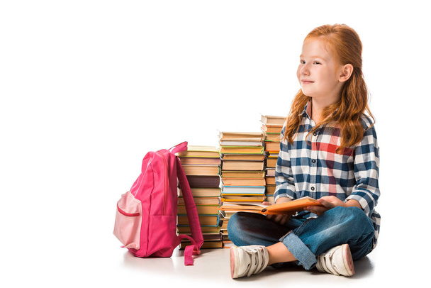 positive redhead schoolkid sitting near books and pink backpack on white  - Foto, immagini