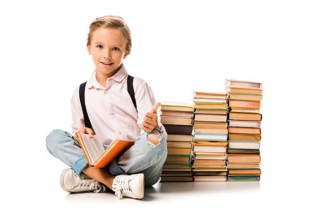 cheerful kid sitting with crossed legs near books and showing thumb up on white  - Фото, изображение