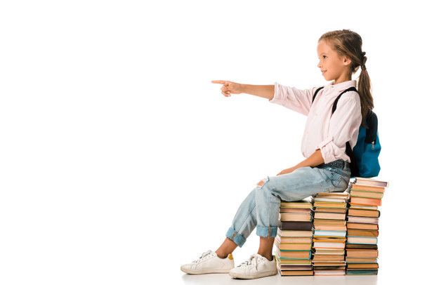 cheerful kid sitting on books and pointing with finger on white  - Valokuva, kuva