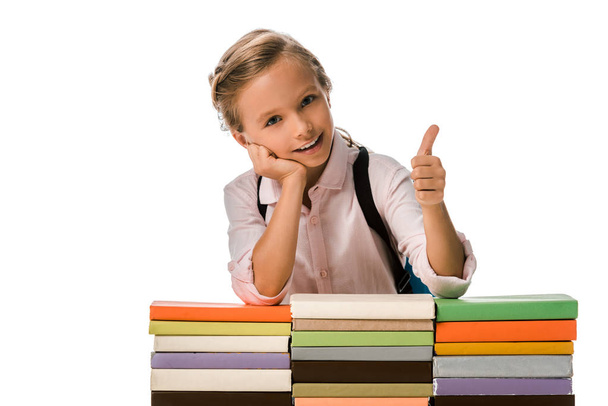 χαρούμενο σχολιαρόπαιδο που δείχνει τον αντίχειρα κοντά σε πολύχρωμα βιβλία απομονωμένα στα λευκά  - Φωτογραφία, εικόνα
