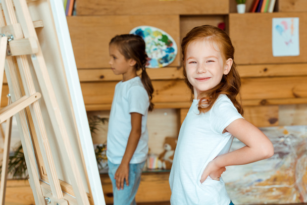 美術学校の子供の近くのヒップに手を携えて立つ幸せな赤毛の子供の選択的な焦点  - 写真・画像