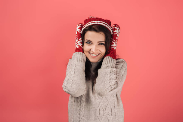 cheerful woman in knitted hat and gloves covering ears on pink  - Фото, изображение