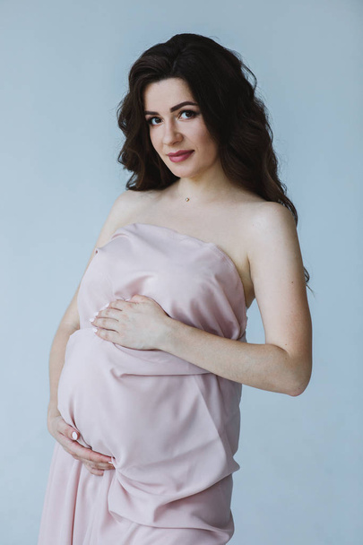 Cute young mother with long wave hair, natural make up looking at camera and holding her belly. Woman podinh in isolated studio and waiting for baby. Concept of love and motherhood. - Zdjęcie, obraz