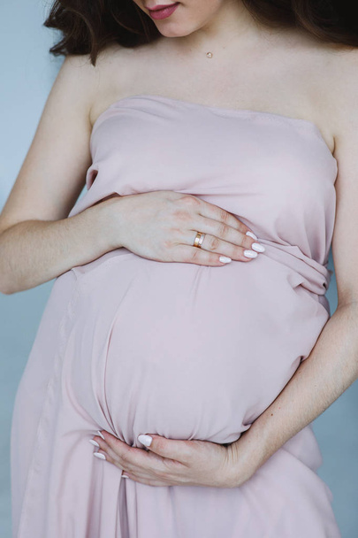 Zbliżenie kobiety w ciąży trzymającej się za brzuch. Anonimowa kobieta w różowej sukience czeka na dziecko. Pojęcie macierzyństwa i opieki. - Zdjęcie, obraz
