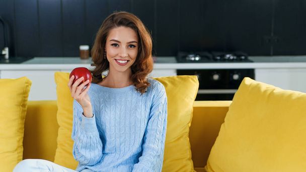 Beautiful female vegetarian in casual clothes holds fresh red apple while sitting on comfortable yellow sofa in the stylish modern kitchen. - Fotoğraf, Görsel