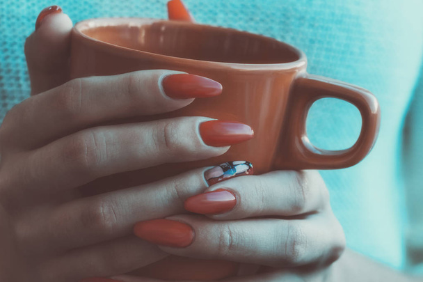 νεαρή κοπέλα με κόκκινο μανικιούρ κρατώντας ένα πορτοκαλί φλιτζάνι αρωματικό τσάι, έννοια του φθινοπώρου. Χειμερινή ιδέα. Κλείσε. Γραμμένο - Φωτογραφία, εικόνα