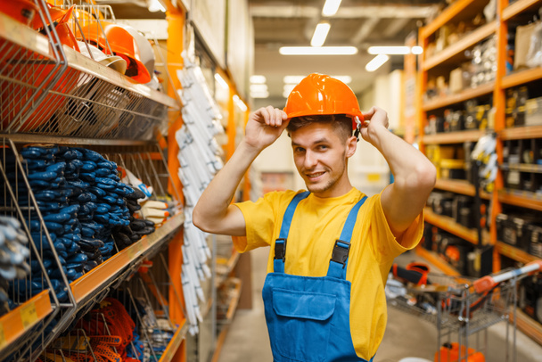 Construtor masculino tentando capacete na prateleira na loja de ferragens. Construtor em uniforme olhar para as mercadorias em loja diy
 - Foto, Imagem
