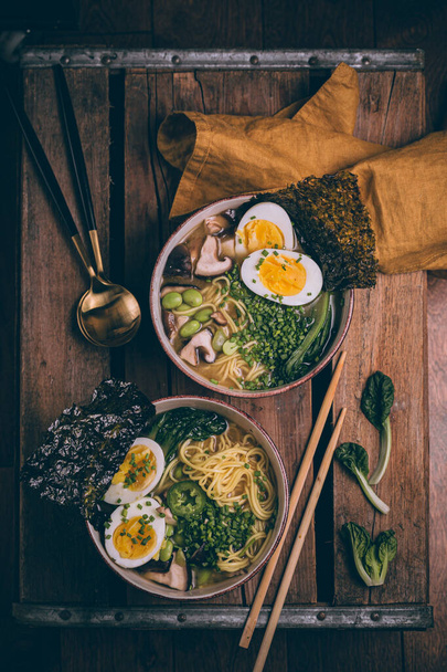 Γιαπωνέζικη σούπα Ράμεν. Παραδοσιακή Ιαπωνική κουζίνα. Ασιατικός ζωμός με noodles, bok choy, μανιτάρια σιτάκι, edamame. Ρουστίκ σκοτεινή διάθεση. Βραστό αυγό σε σούπα. Dashi ζωμός. Ελικόπτερα, κουτάλια σε ξύλινο τραπέζι. - Φωτογραφία, εικόνα