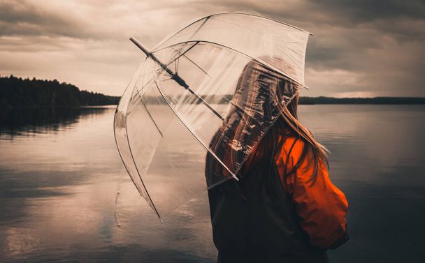 Girl with an umbrella on the lake in a gray-orange jacket, cloudy weather or dark theme - Fotoğraf, Görsel