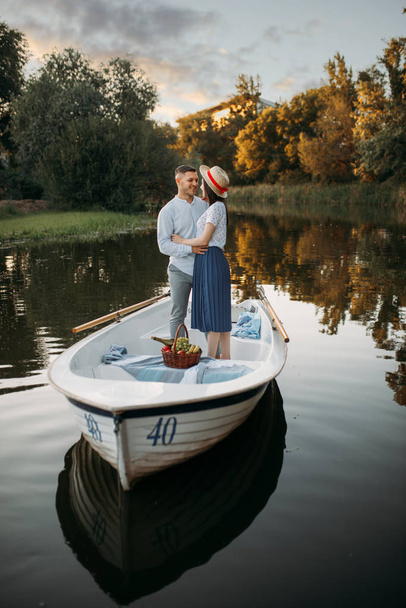 Love couple standing in a boat on quiet lake at summer day on sunset. Romantic meeting, boating trip, man and woman walking along the lake - Photo, Image